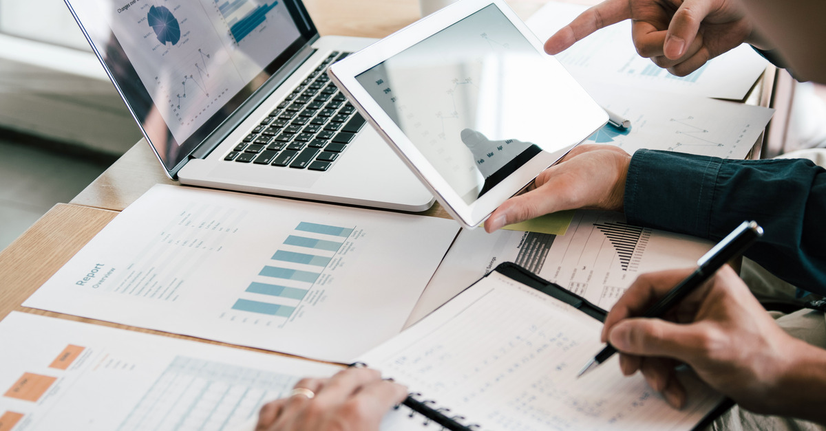 A close-up of business coworkers using a tablet and laptop to plan their business report. There are also scattered papers.